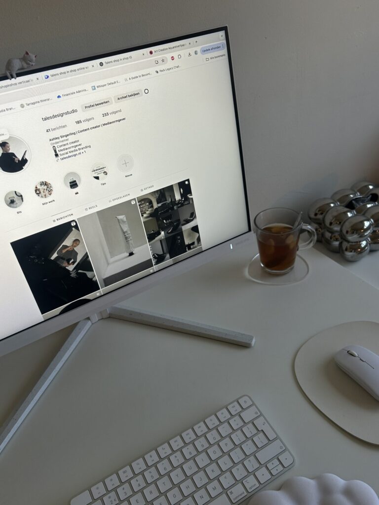 a photo of a desk with a monitor, cup of tea, mouse, and keyobard. It's a white desk and a white wall behind it. On the screen, there is an Instagram Feed open.