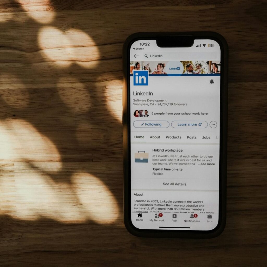 LinkedIn content framework. A smartphone displays the LinkedIn profile page on a wooden surface, with soft, dappled sunlight creating patterns. The atmosphere is calm and professional.